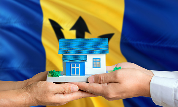 two hands exchanging a small model house set against a background of the Barbados flag representing home ownership and investment 9 ways to improve housing options